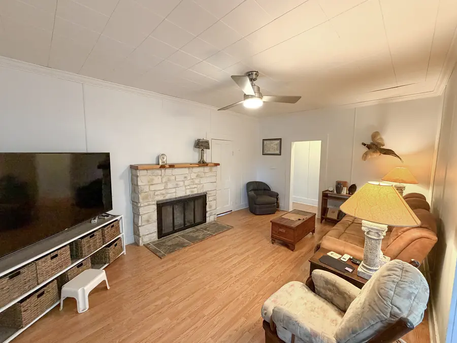 Living area with a stone fireplace, television console, brown sofa, armchairs, lamps, and a lighted ceiling fan over wood-look flooring. Bond Ranch ranch house interior.